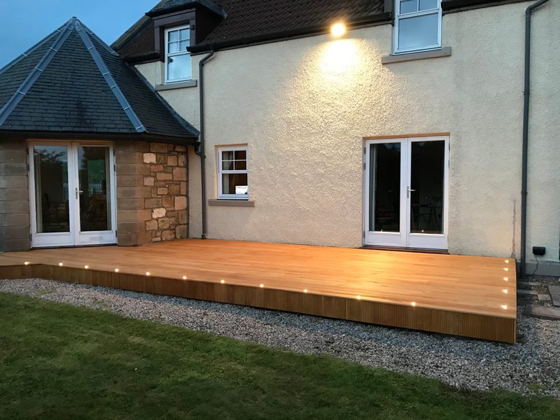 Wooden deck extending from a beige house with glass doors, a small window, a stone wall on the left, gravel border, and lawn; LED edge lights.
