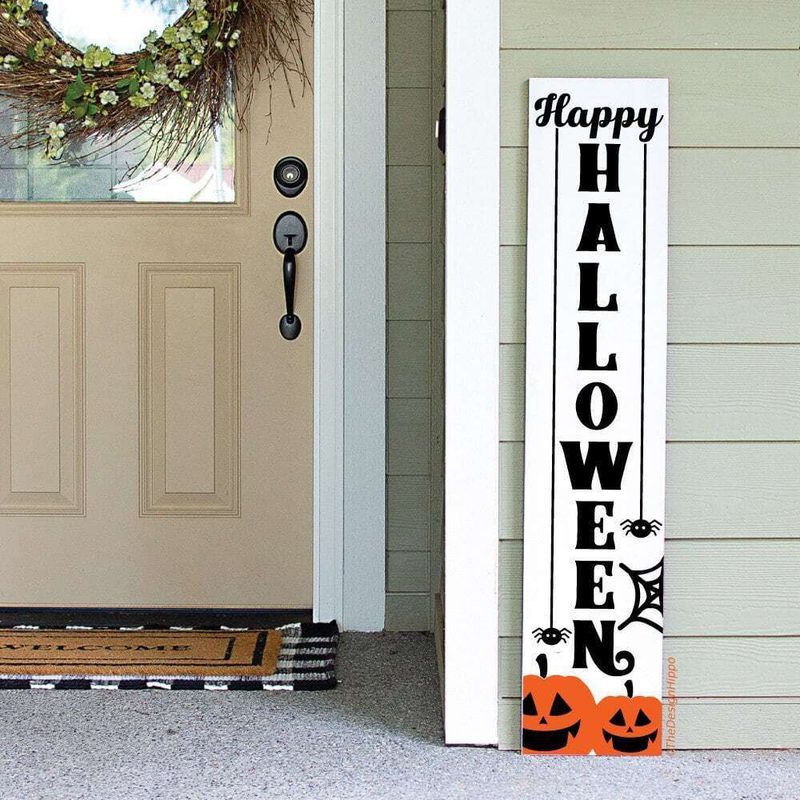 Beige front door with a wreath and black handle; a vertical white "Happy Halloween" sign leans against light green siding, with orange pumpkins at the base.