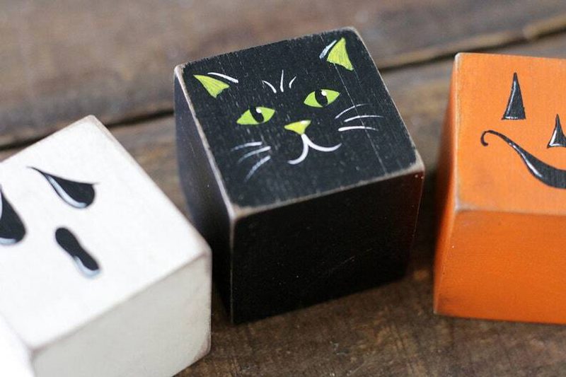 Three painted wooden cubes on a wooden surface: white cube with black eyes, black cube with a green-eyed cat, orange cube with jack-o-lantern grin.