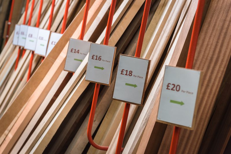 Row of wooden planks in varying brown tones on orange supports, with white price signs (£14–£20 per piece) and green arrows.