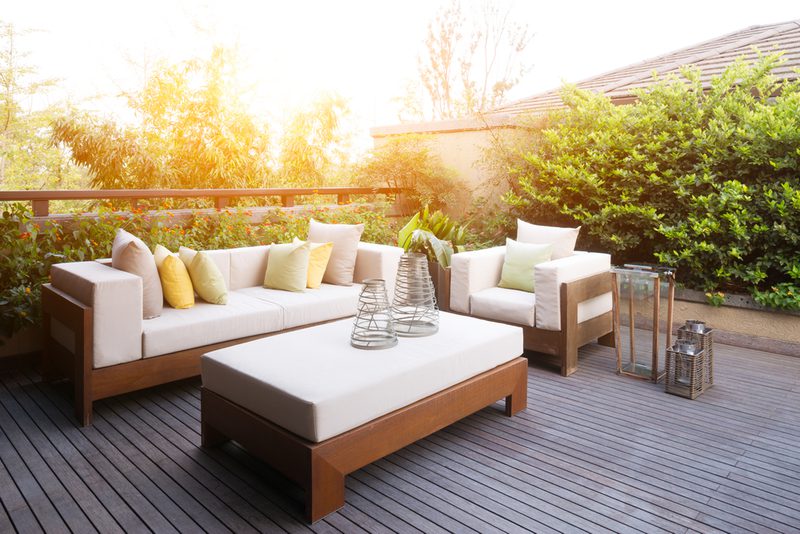 Sunlit rooftop deck with beige cushioned wooden sofa and chair, a white-cushioned coffee table, metal lanterns, pastel pillows, and a green hedge backdrop.