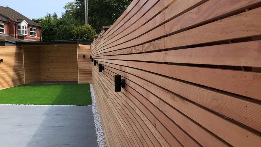 Long horizontal wooden fence along a modern garden, with black wall lights, gray paving, a white gravel border, and a small green lawn.