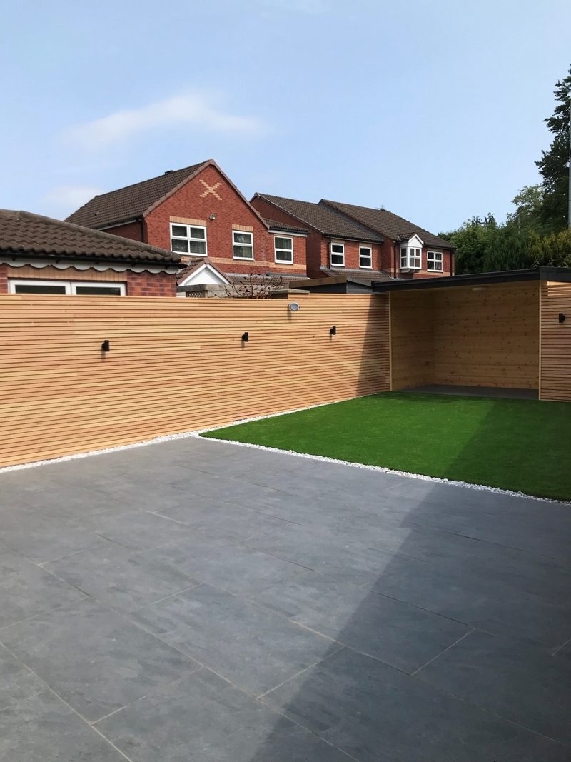 Modern backyard with gray stone patio, wooden privacy fence, patch of artificial grass, white pebble border, and red-brick houses against a blue sky.