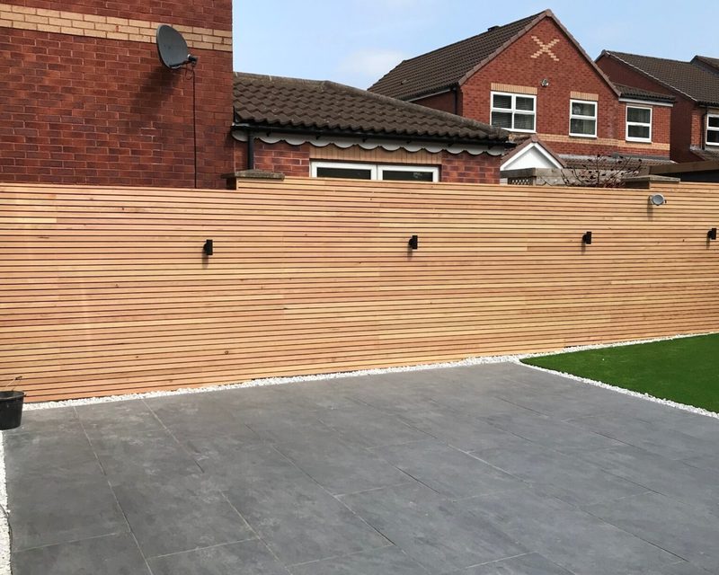 Backyard with a tall wooden fence, grey tiled patio, white gravel border, and artificial grass; brick houses and a satellite dish visible.