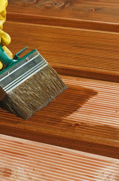 A yellow-gloved hand holds a wide paintbrush and stains wooden deck boards, with sunlight casting shadows across the grooved planks.