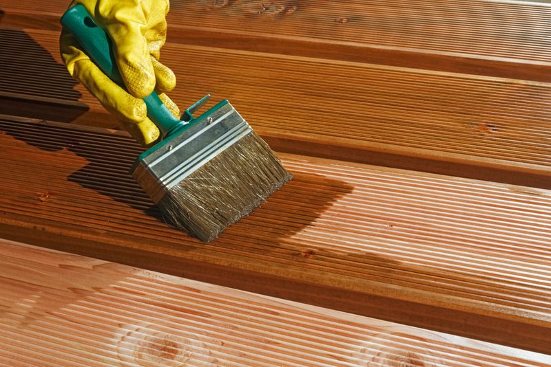 A yellow-gloved hand holds a wide paintbrush and stains wooden deck boards, with sunlight casting shadows across the grooved planks.