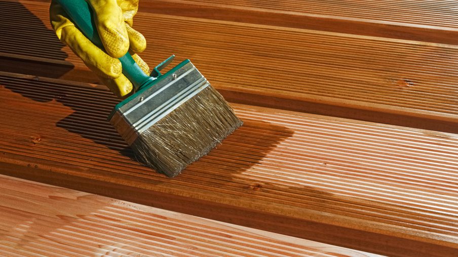 A yellow-gloved hand holds a wide paintbrush and stains wooden deck boards, with sunlight casting shadows across the grooved planks.