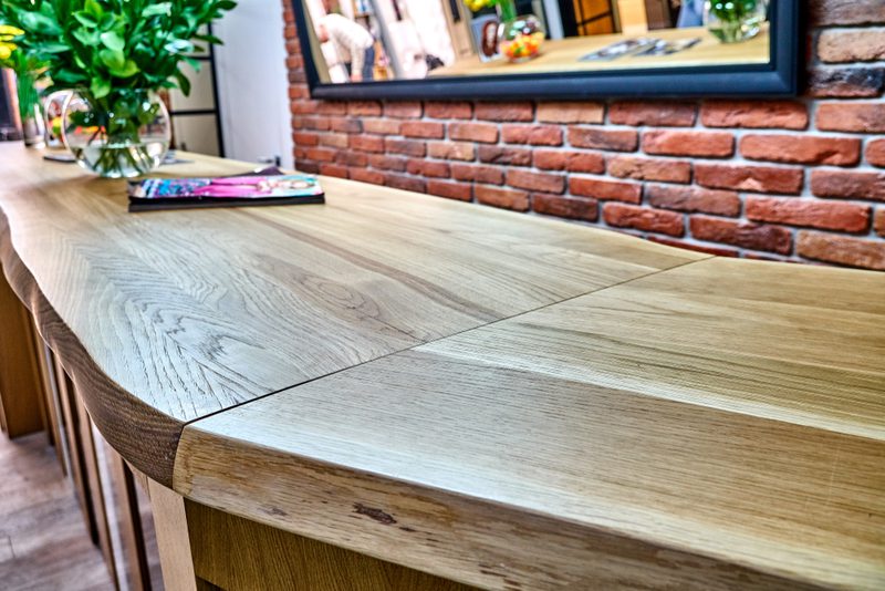 Close-up of a curved, light-wood kitchen island with grain, against a red brick wall; a mirror, a potted plant, and magazines rest on the counter.