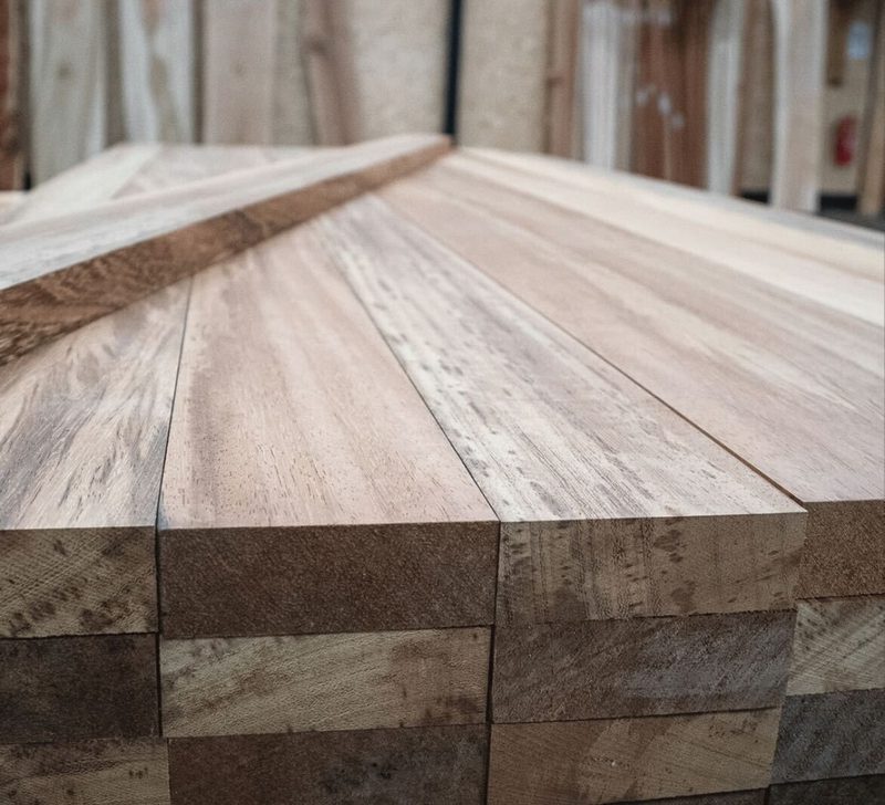 Stacked light-to-medium brown wooden planks with visible grain, arranged in a workshop; perspective view along the length of the boards.