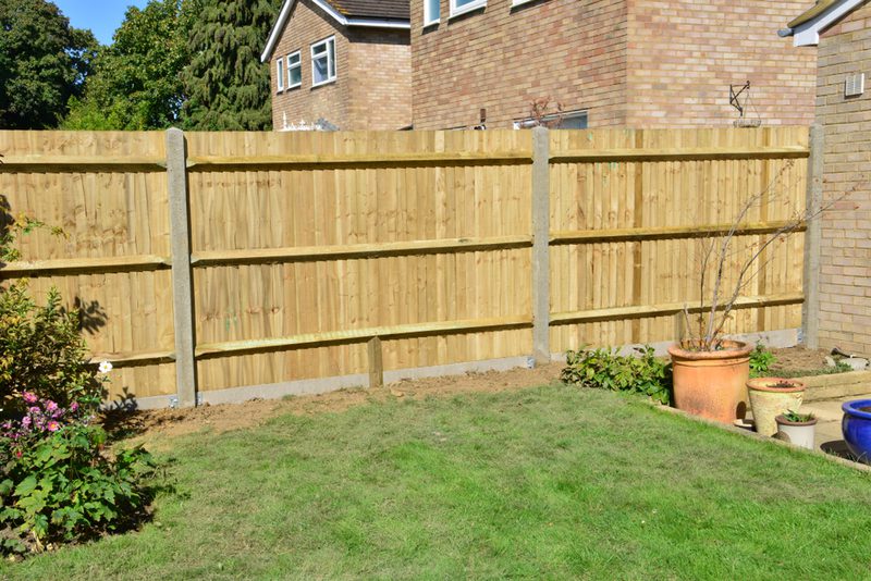 Backyard with a tall wooden fence and concrete posts, a green lawn, and several potted plants beside a brick house; blue sky and trees in the background.