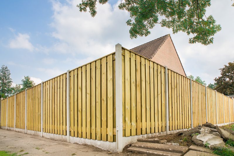 Long wooden privacy fence with vertical slats and concrete posts runs along a sidewalk; a brick roof rises behind, with green trees and blue sky.