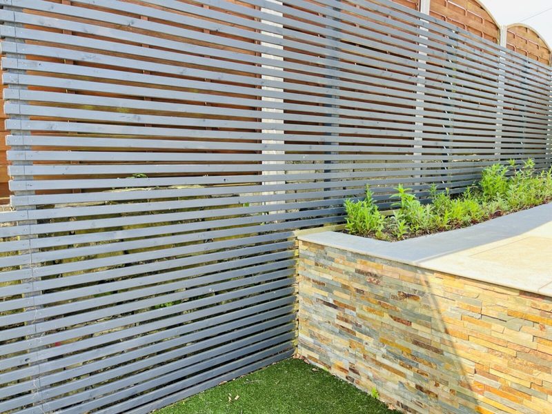 Gray horizontal slat fence beside a stacked-stone planter with small green plants, a narrow lawn at the base, sunlit with soft shadows.
