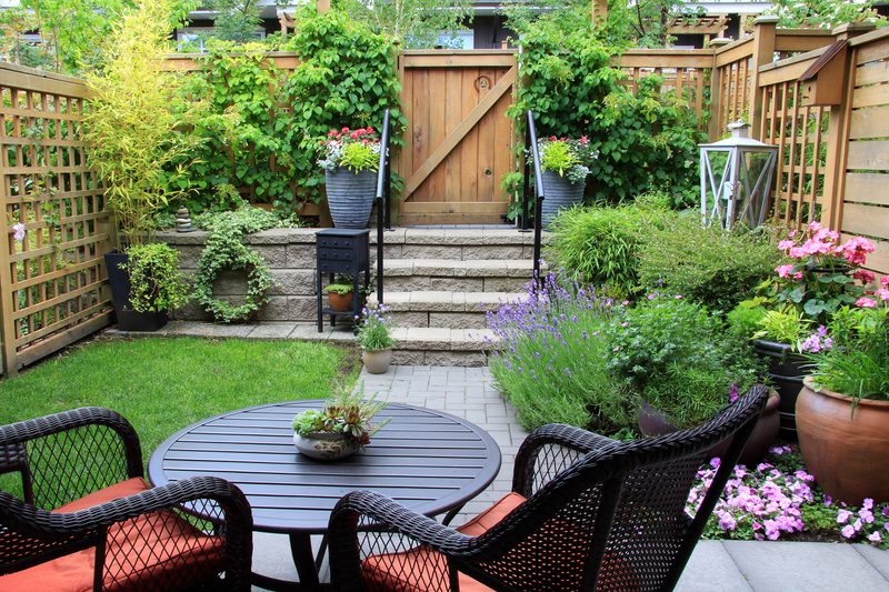 Cozy backyard patio with a round black table and wicker chairs, surrounded by potted flowers, lavender, and lush greenery, stone steps and a wooden gate.