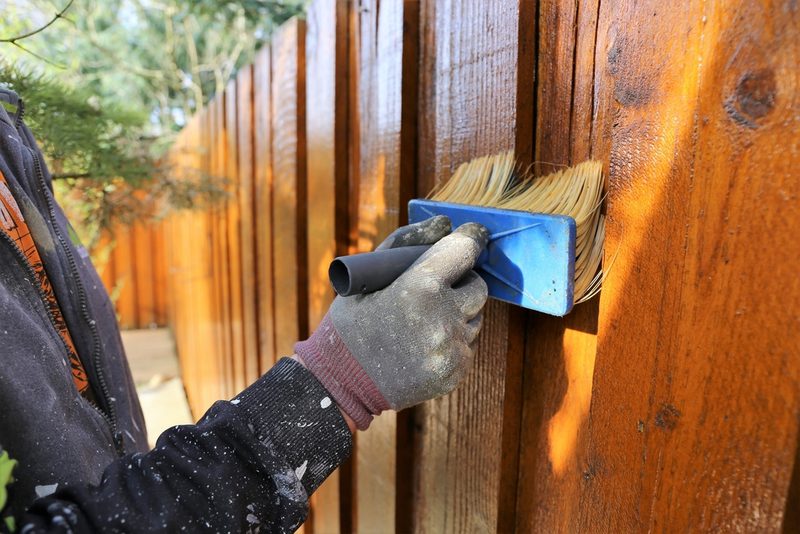 Person wearing a paint-splattered glove uses a blue brush with light bristles to stain an orange-brown wooden fence outdoors.