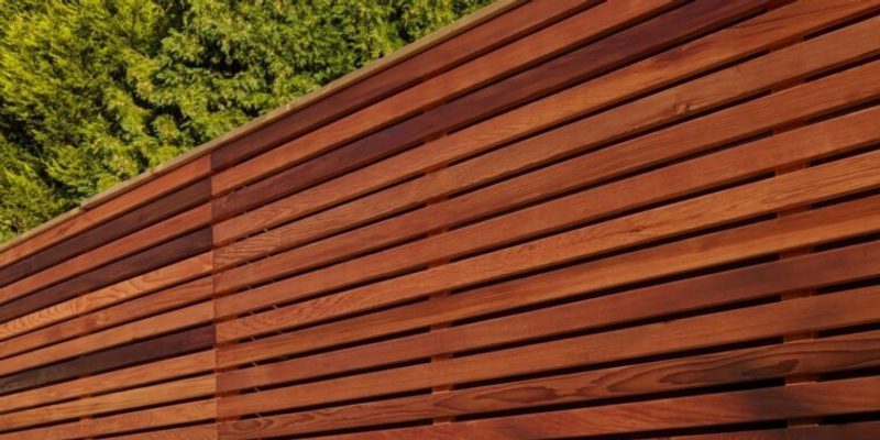 Reddish-brown horizontal wooden slats forming a wall or fence, with green trees visible in the upper left corner.