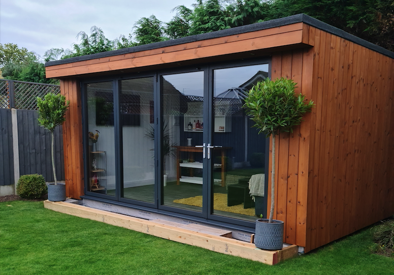 Wood-clad garden room with large glass doors and dark frames on a wooden deck; two potted topiary trees flank the entrance, green lawn around.