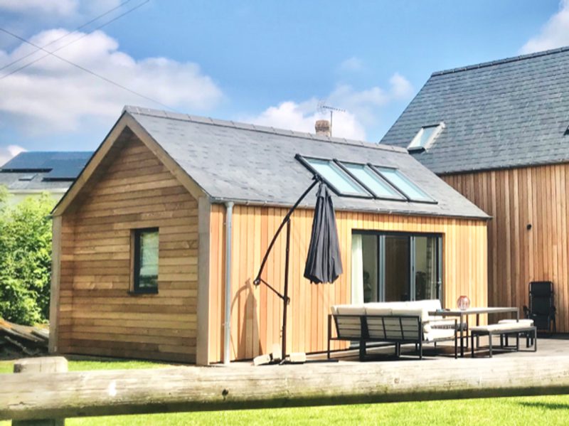 A wooden cabin with a sloped roof and skylights sits on a sunny day, with a deck and outdoor furniture including a sofa, chairs, a table, and a closed umbrella.