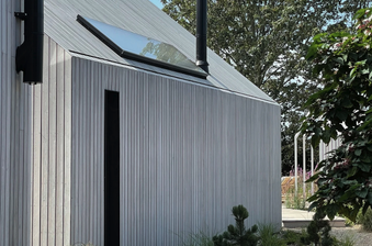 A modern corrugated metal building with a sloped roof and skylight; a narrow dark doorway beside a gravel path and small plants along the edge.