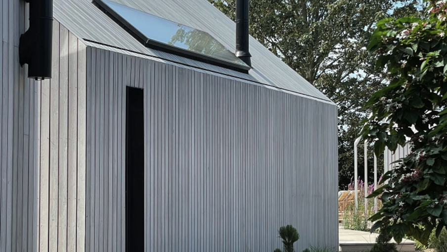 A modern corrugated metal building with a sloped roof and skylight; a narrow dark doorway beside a gravel path and small plants along the edge.