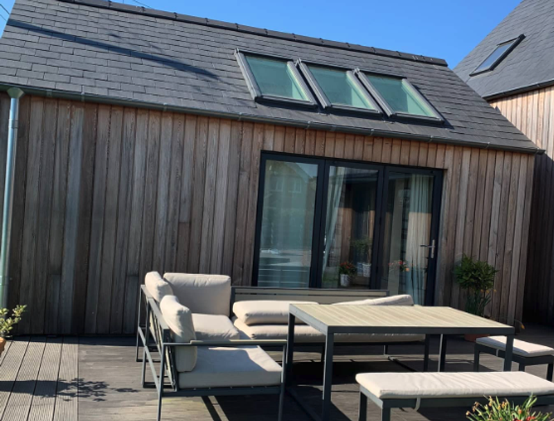 Wood-clad house with a sloped roof, three skylights, and glass doors, featuring a beige-cushioned outdoor sofa set and rectangular table on a wooden deck.