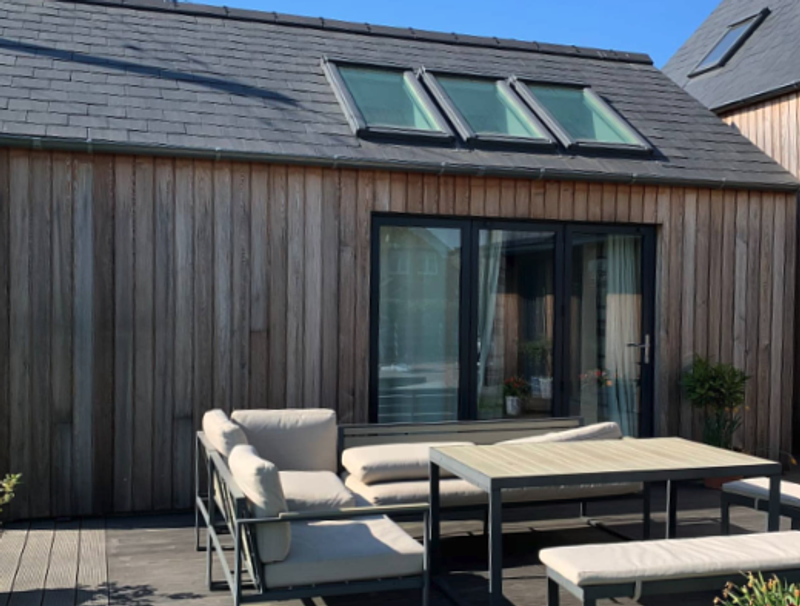 Wood-clad house with a sloped roof, three skylights, and glass doors, featuring a beige-cushioned outdoor sofa set and rectangular table on a wooden deck.