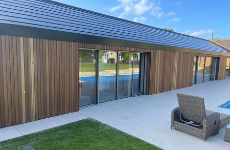 Modern single-story building with wooden siding and large sliding glass doors, a tiled patio, a blue pool beyond, and wicker chairs on a sunny day.