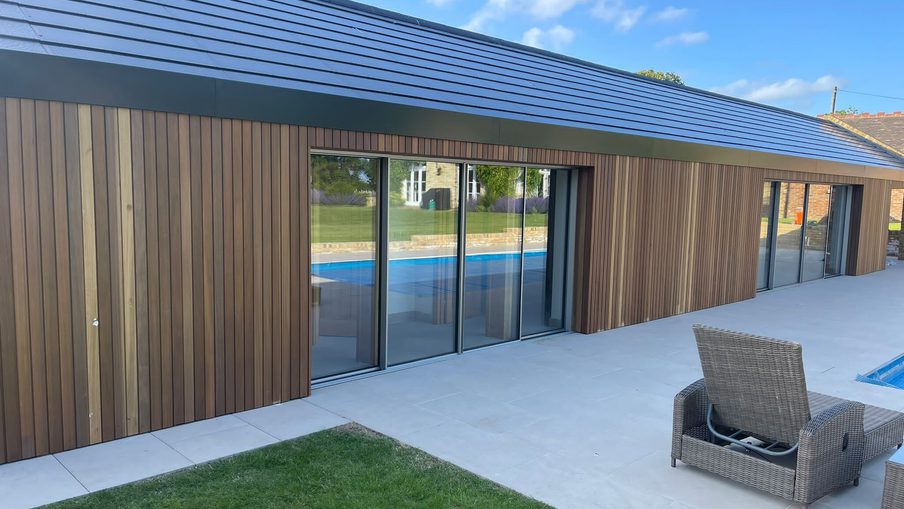 Modern single-story building with wooden siding and large sliding glass doors, a tiled patio, a blue pool beyond, and wicker chairs on a sunny day.