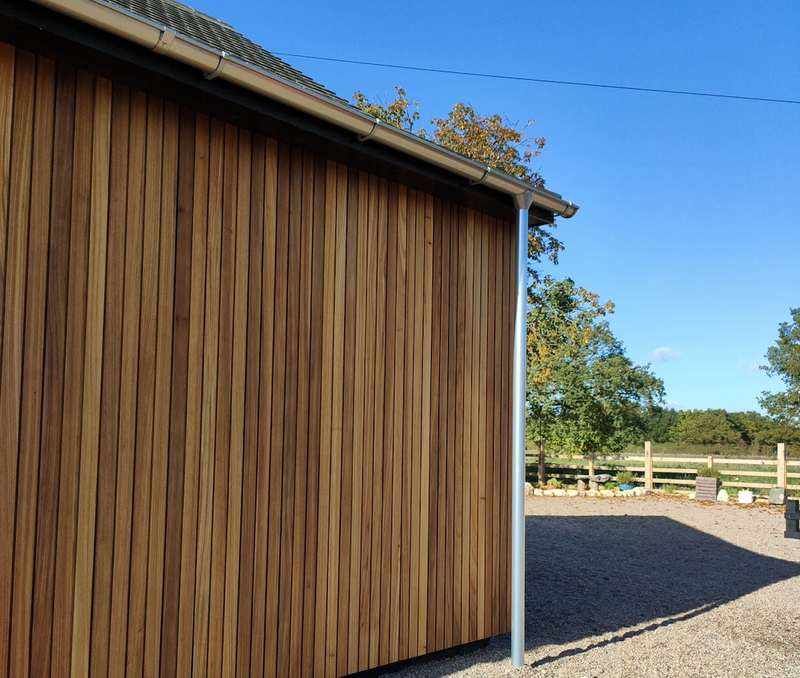 Wooden building with tall vertical planks, a metal downspout on the right, gravel yard, distant fence and trees under a bright blue sky.