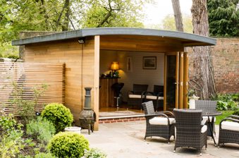 Wooden garden studio with a roof opens to a seating area of wicker chairs; inside a small bar, lamp, and wall art amid greenery and a brick wall.
