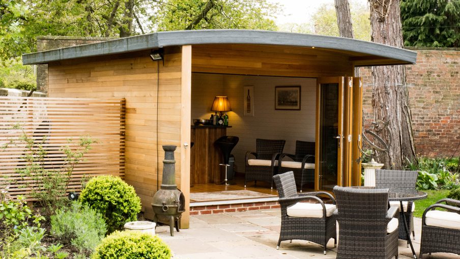 Wooden garden studio with a roof opens to a seating area of wicker chairs; inside a small bar, lamp, and wall art amid greenery and a brick wall.