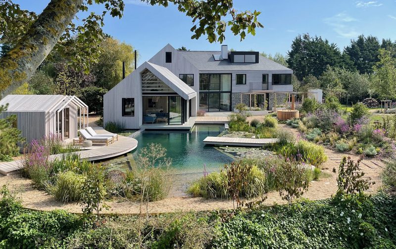 Modern gray house with large glass walls and a triangular pavilion, beside a pool with a wooden deck, set in a lush landscaped garden on a sunny day.