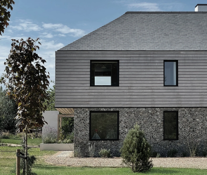 Two-story house with a stone base and gray wooden upper level, a sloped roof, dark-framed windows, and a gravel yard with shrubs and a small tree.