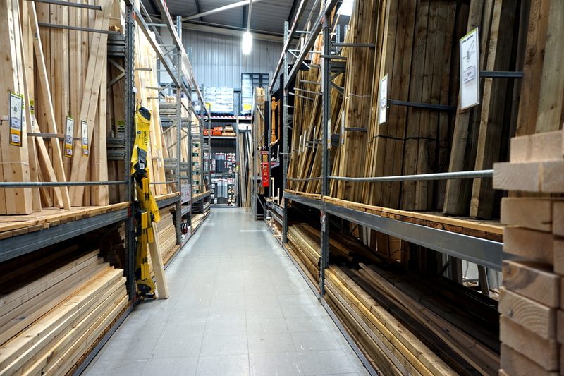 Wide warehouse aisle with tall metal racks on both sides, stacked lumber and planks, signs on shelves, under bright overhead lighting.