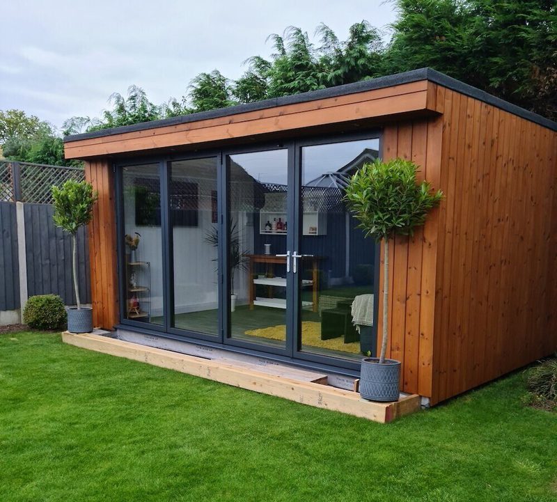 A wooden garden room with large glass sliding doors, surrounded by a green lawn and a wooden deck; two potted topiary trees frame the entrance.