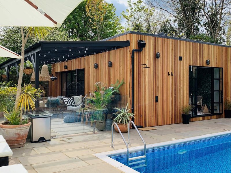 Wood-clad poolside retreat with glass doors, string lights, and a cozy outdoor lounge; potted plants line the patio beside a blue-tiled swimming pool.