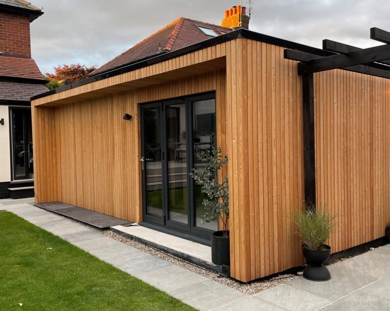 Modern wooden extension with vertical timber cladding, large black-framed glass doors, a concrete path, and potted plants by the doorway under an overcast sky.