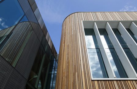 Two modern facades meet: a curved wood-clad wall with vertical slats and white-framed glass windows, beside a dark glass surface under a blue sky.