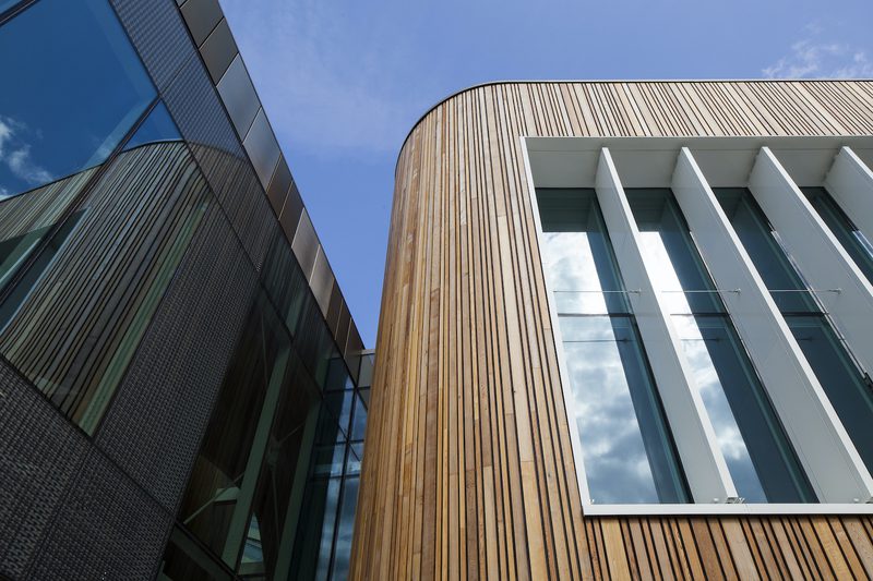 Two modern facades meet: a curved wood-clad wall with vertical slats and white-framed glass windows, beside a dark glass surface under a blue sky.