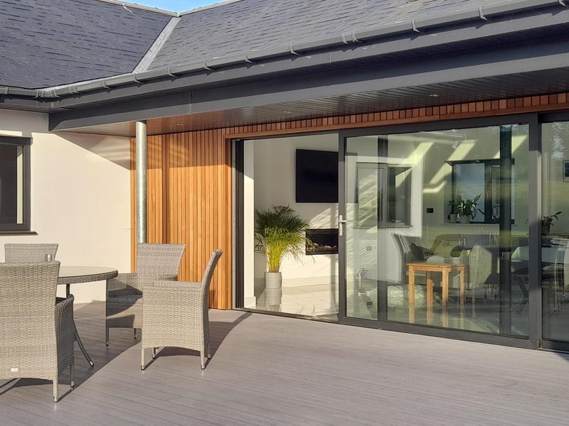 Modern patio with gray decking, a wicker dining set, and large glass sliding doors opening to a bright interior with potted plants.