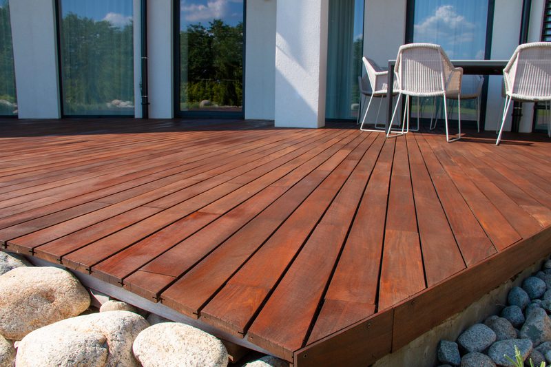 Red-brown wooden deck attached to a modern glass-walled home, with white wicker chairs around a table; large stones beneath.