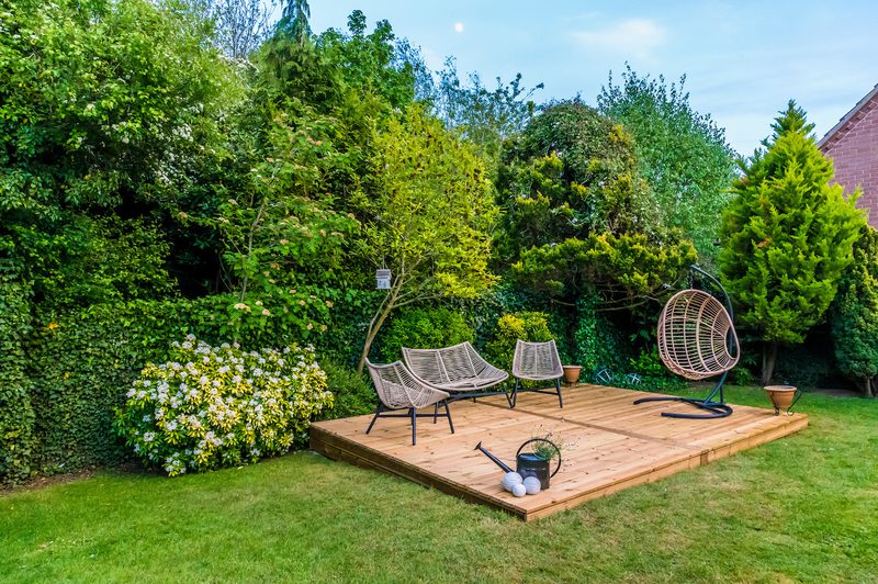 Wooden garden deck with woven lounge chairs and a circular hanging chair, surrounded by green hedges and trees on a sunny lawn.