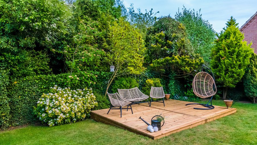 Wooden garden deck with woven lounge chairs and a circular hanging chair, surrounded by green hedges and trees on a sunny lawn.