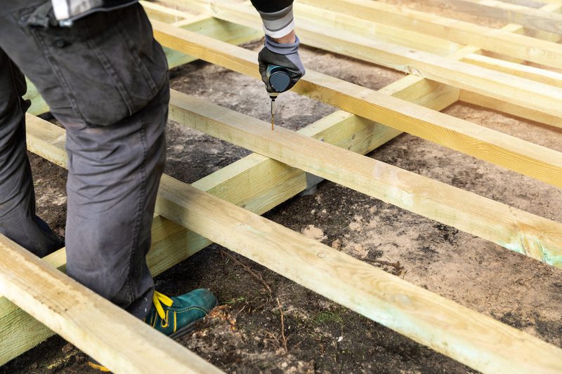 A person in gloves and dark pants drills a screw into a wooden frame at a construction site.