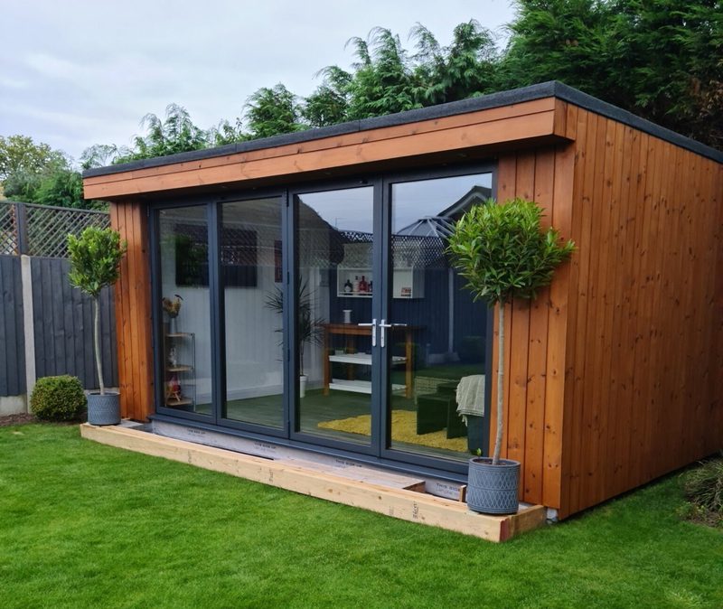 Wooden garden studio with large glass doors, two potted topiary trees, and a wooden deck on a green lawn.