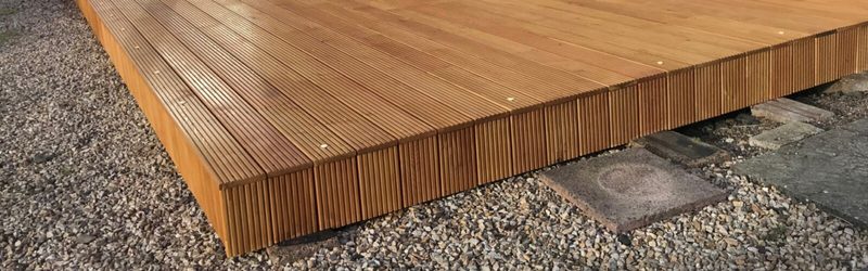 Low-angle view of a warm brown wooden deck with narrow grooved boards, sitting on gravel with stepping stones nearby.