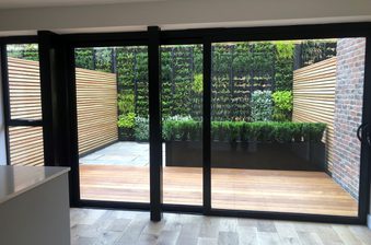 Room with large black-framed glass doors opening to a wooden deck and lush garden: vertical plant wall, trimmed hedge, wooden screens, brick wall.