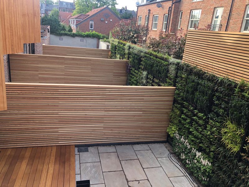 Patio with layered horizontal wooden privacy screens, a stone-paved ground, and a green hedge wall along a brick building backdrop.