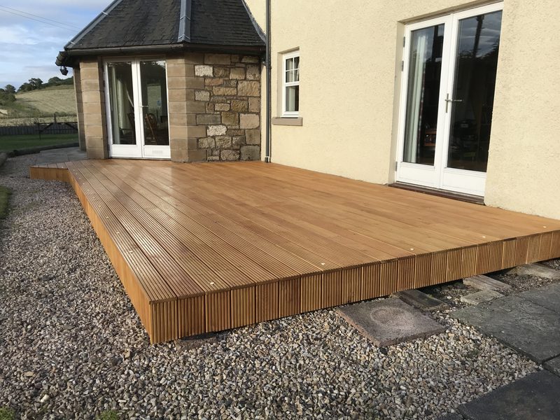New wooden deck attached to a cream-colored house, featuring grooved boards and a vertical slat edge, extending over gravel with glass doors.