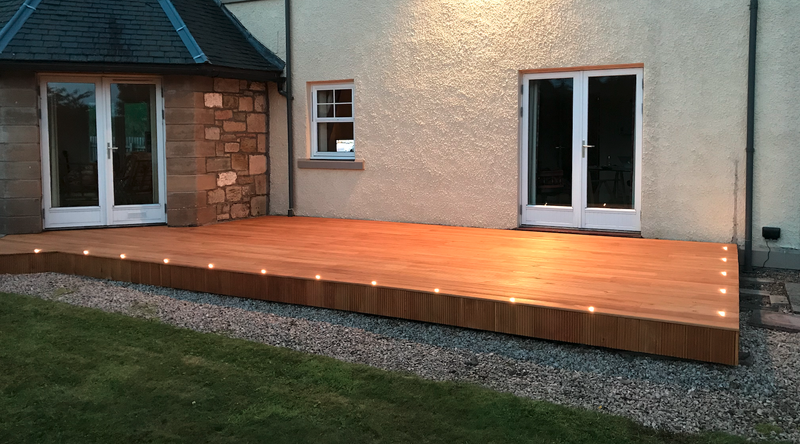 Wooden deck along the back of a house with small edge lights; stone corner, white-framed window and double doors on a stucco wall, gravel and lawn.