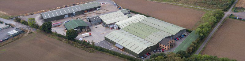 Aerial view of a business park with several long warehouses featuring pale green and white roofs, surrounded by farmland, roads, and parked cars along the edges.